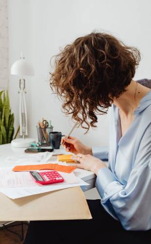 A woman in a blue blouse writing at a desk with a calculator and documents.