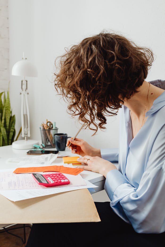 about-us-02 A woman in a blue blouse writing at a desk with a calculator and documents.
